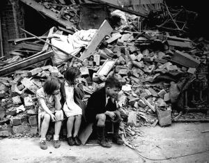 Children of an eastern suburb of London, who have been made homeless by the random bombs of the Nazi night raiders, waiting outside the wreckage of what was their home